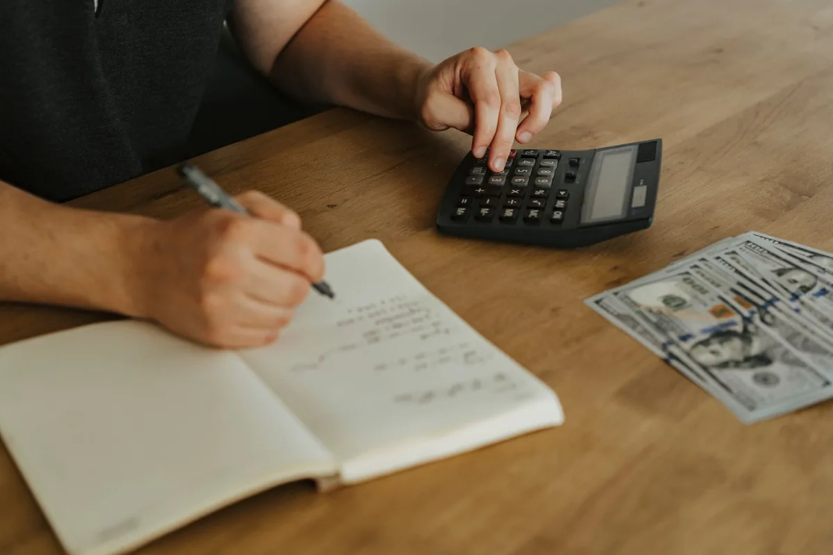 A person writing figures in an open notebook with one hand and pressing a key on a black desk calculator with the other, with a fan of banknotes lying on the wooden table