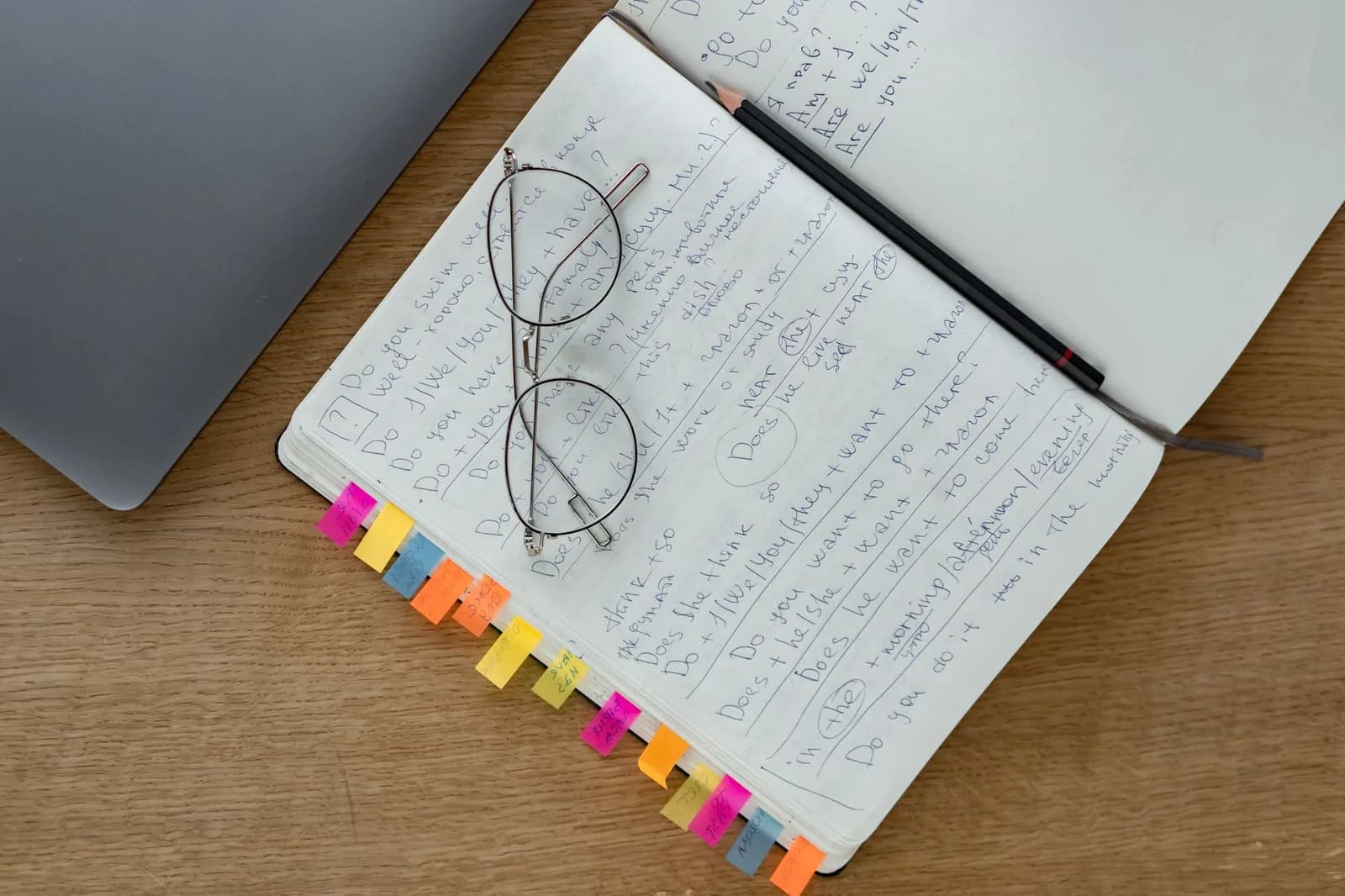 An open notebook full of handwritten notes with a row of coloured sticky tabs along its edge, a pair of round glasses and a pencil resting on the page, beside a closed laptop on a wooden desk