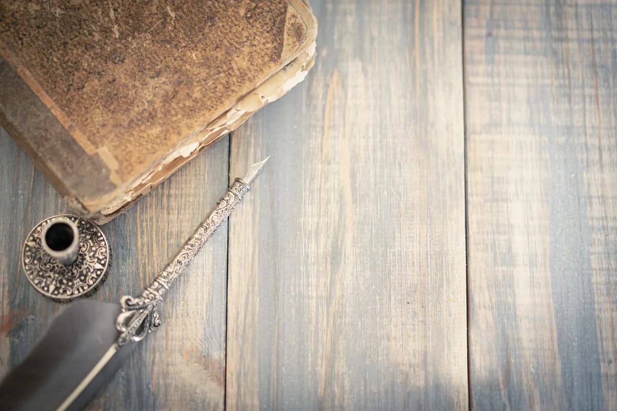 A thick, worn leather-bound book with frayed corners lying on weathered wooden boards beside an ornate silver dip pen and a small metal inkwell