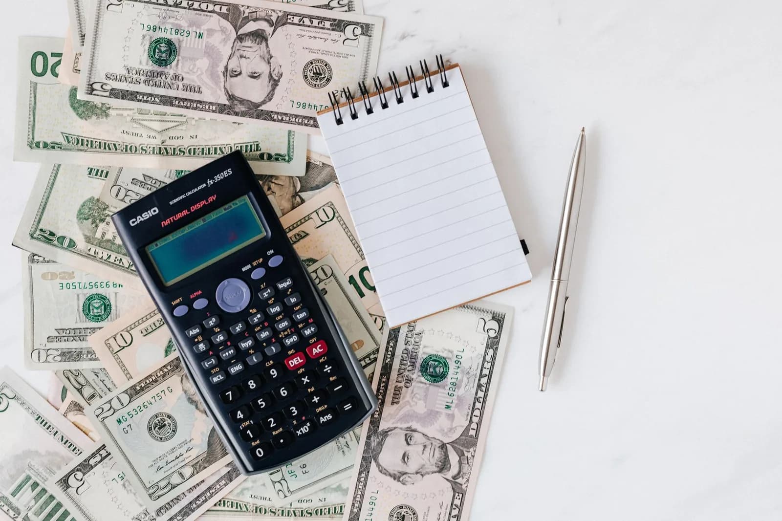 Overhead view of a scientific calculator and a blank spiral notepad resting on a fan of US dollar bills, with a silver pen laid beside them on a white marble surface