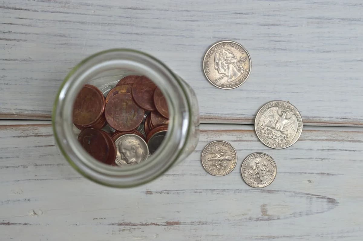 Overhead view of a glass jar half full of pennies with a few quarters and dimes beside it on a weathered white wooden table