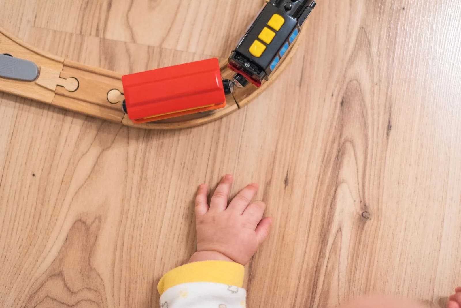 A baby hand resting on a light wooden floor beside a curved wooden toy train track with a red and a black toy train car