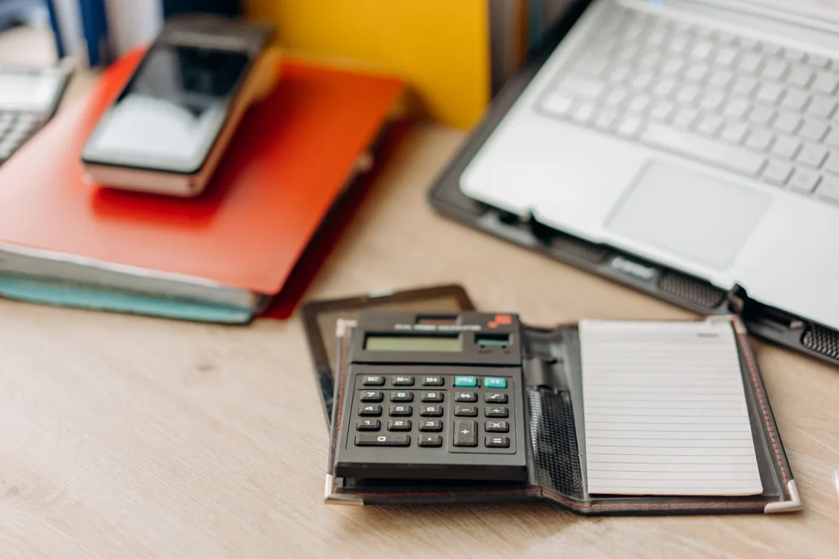 A pocket calculator in a leather folio with a blank lined notepad on a pale wooden desk, beside an open laptop and a stack of red and green folders