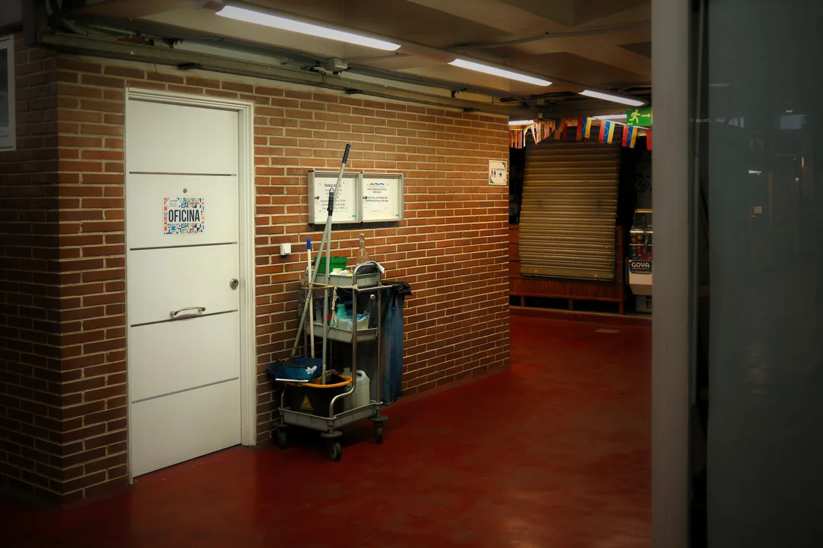 A janitorial cart loaded with a mop, bucket and cleaning supplies parked against a brick wall beside a white office door in an empty hallway