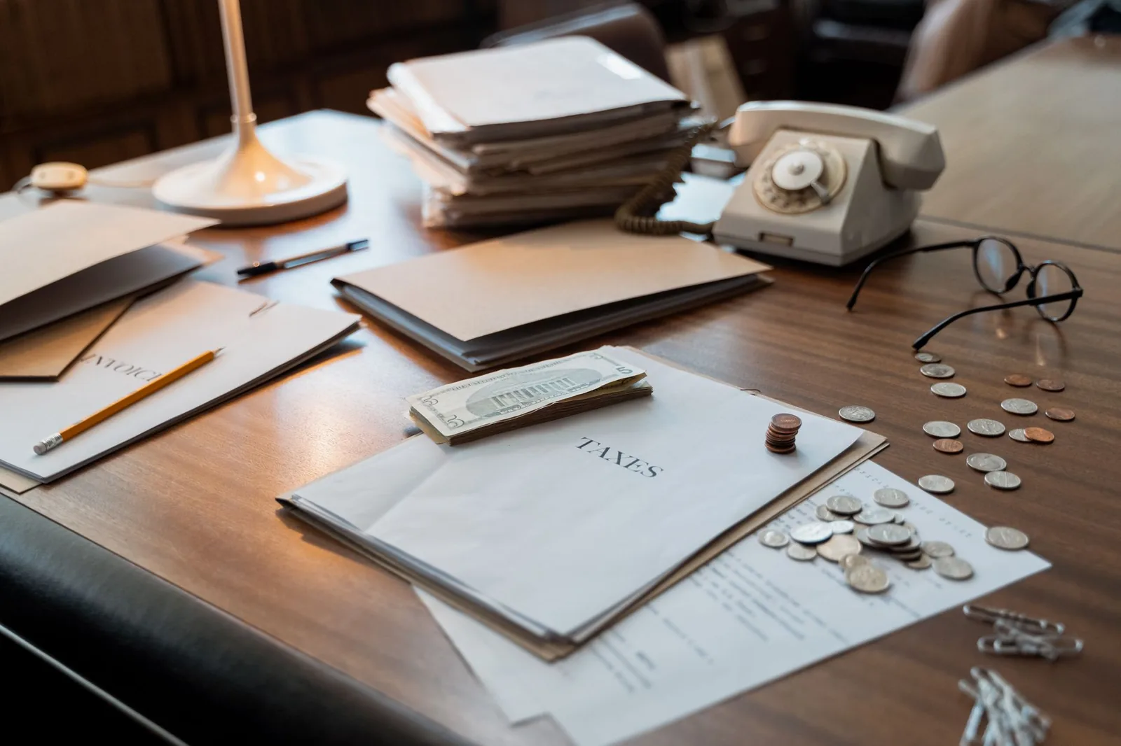 A wooden desk with two folders labeled taxes, a vintage rotary telephone, wire-rimmed reading glasses, loose coins and cash under a warm desk lamp