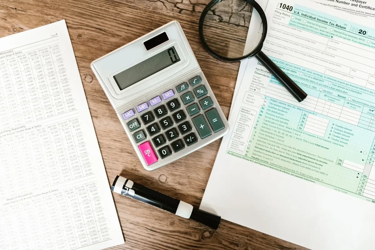 An overhead view of a printed 1040 tax return partly covered by a desktop calculator and a magnifying glass on a wooden table, with a columnar spreadsheet page alongside