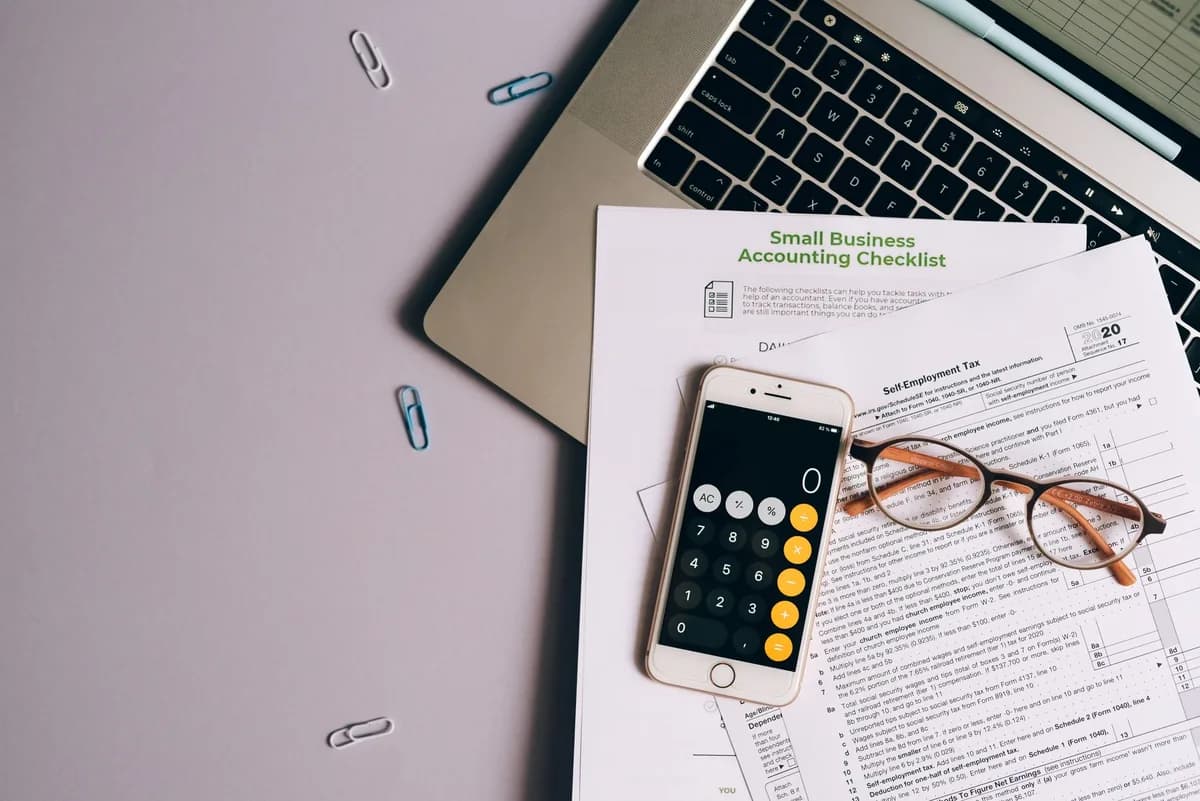 An overhead view of a printed tax form and an accounting checklist on a grey desk, with wire-rimmed reading glasses, a phone showing a calculator app, a laptop and scattered paper clips