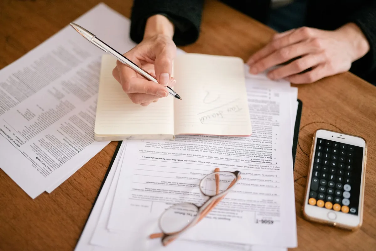 Hands writing in a small notebook on a wooden table covered with printed forms, a pair of reading glasses and a phone showing a calculator