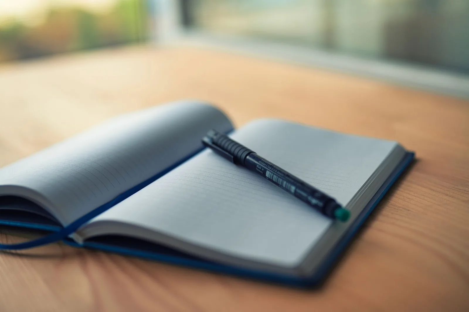 An open lined notebook with a black pen resting across its blank pages on a warm wooden desk by a window
