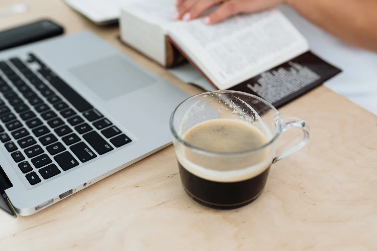 A glass cup of black coffee on a light wooden desk beside an open laptop and a thick open book