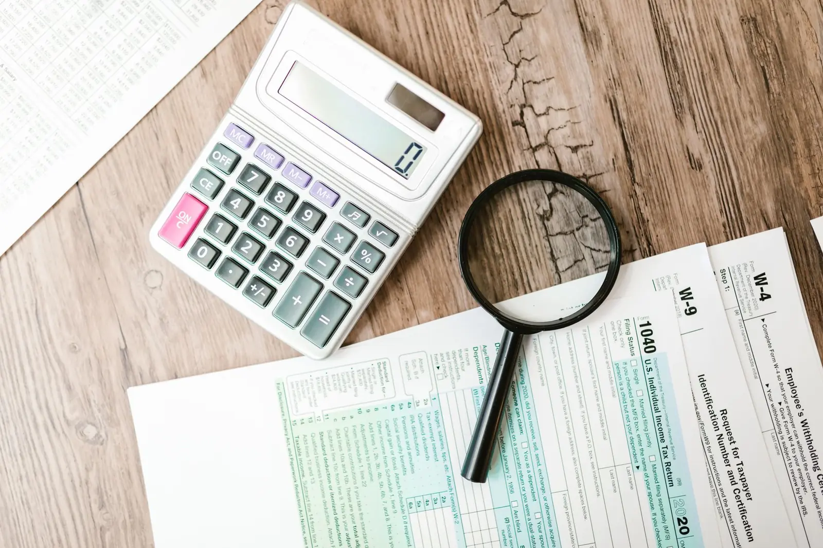 Overhead view of a calculator and a magnifying glass resting on a printed 1040 tax form with W-9 and W-4 forms on a wooden desk