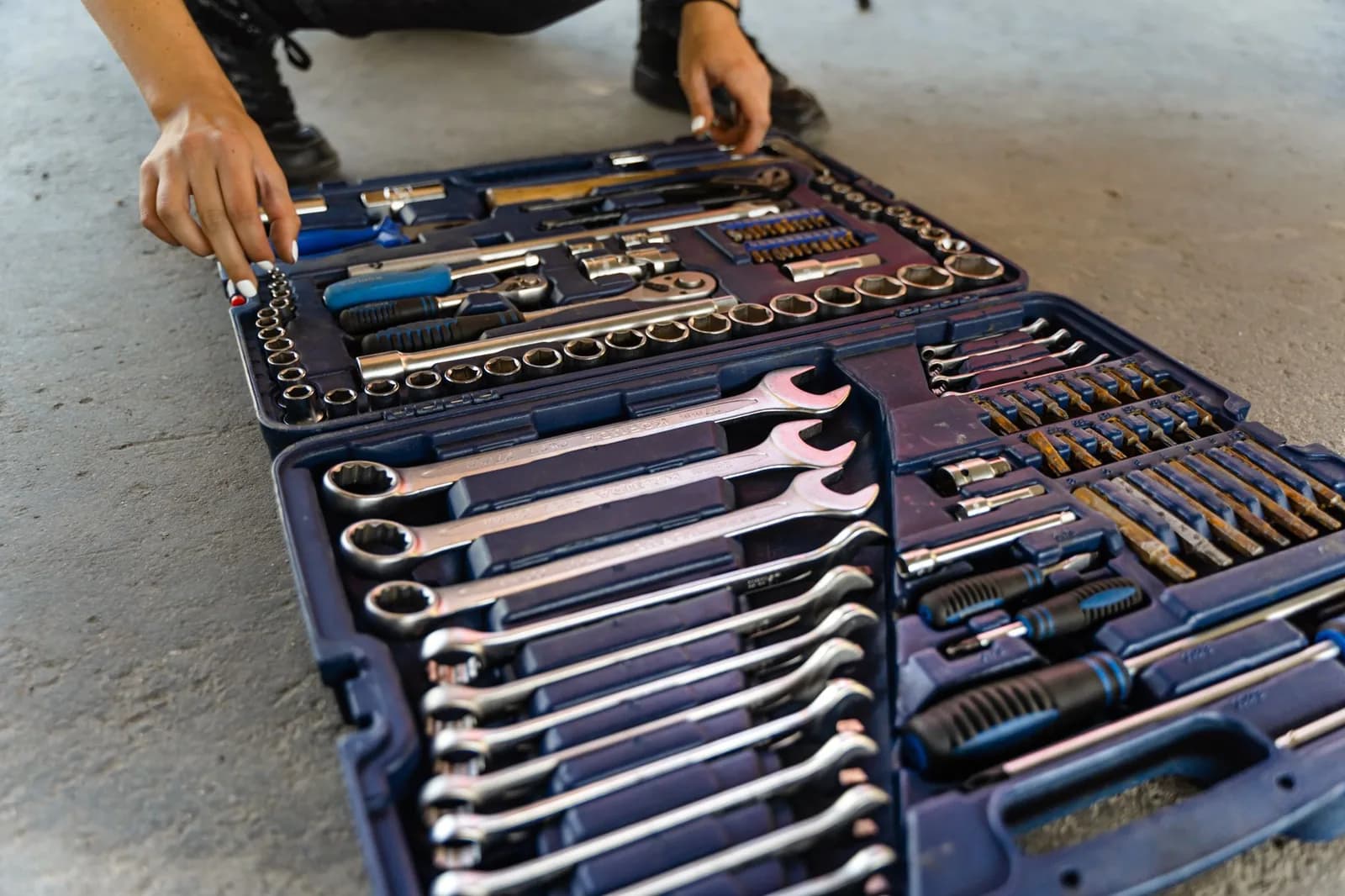 An open blue toolbox on a concrete floor filled with rows of wrenches, sockets and screwdrivers, with a pair of hands reaching for a socket