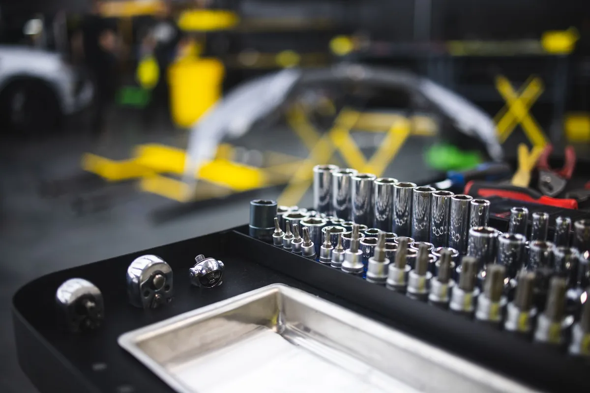 A tray of chrome sockets and ratchet bits in sharp focus on a workbench, with a blurred auto repair shop floor behind it