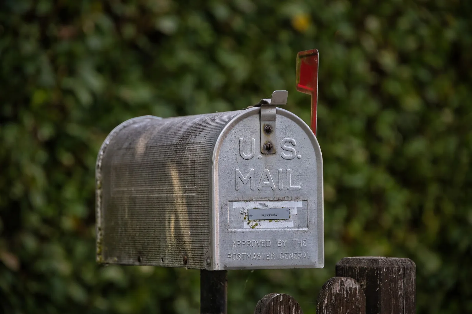A weathered metal mailbox with a raised red flag standing on a wooden post in front of a dark green hedge
