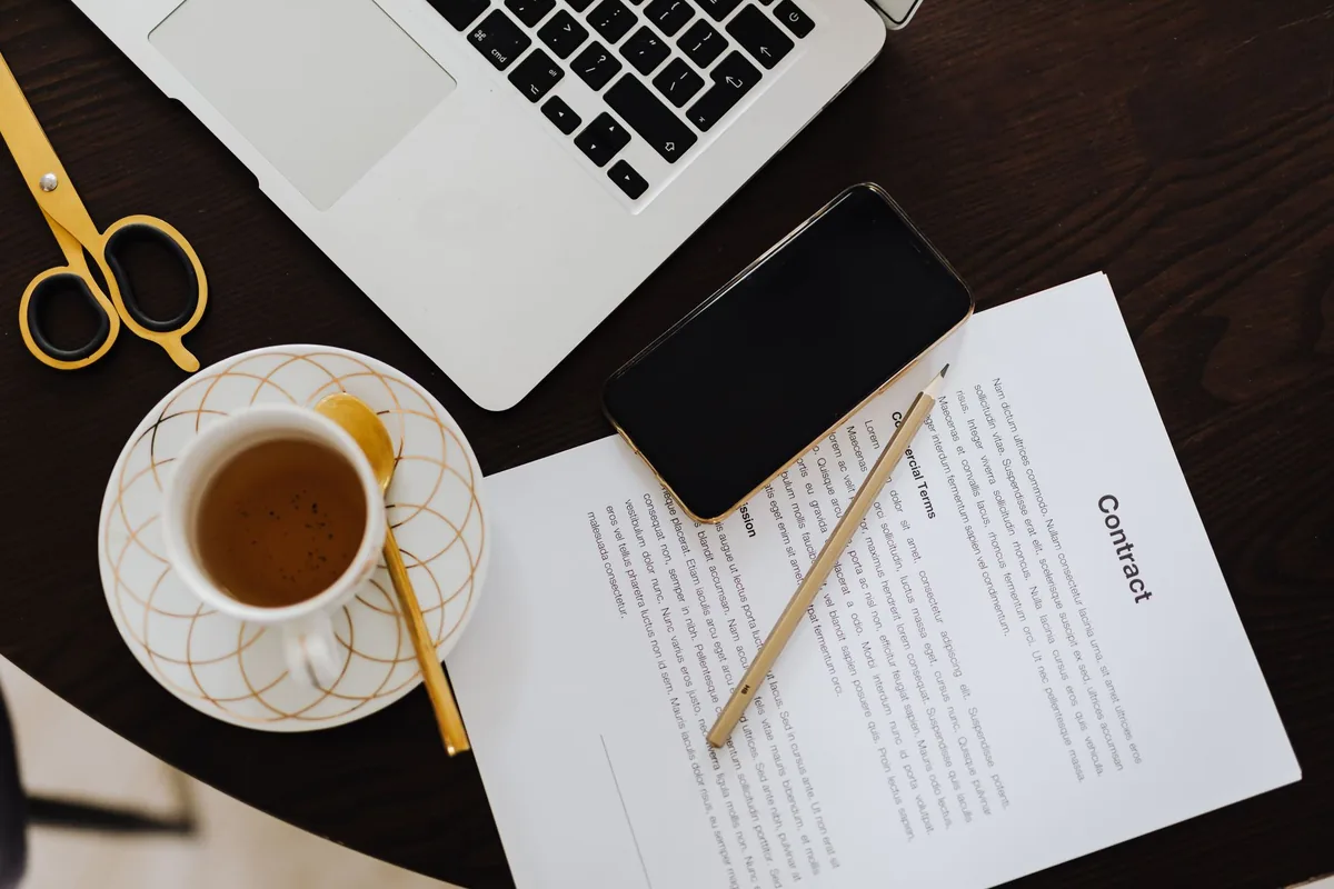 Overhead view of a printed contract with a pencil and a smartphone lying on it, next to a cup of tea, scissors and an open laptop on a dark round table