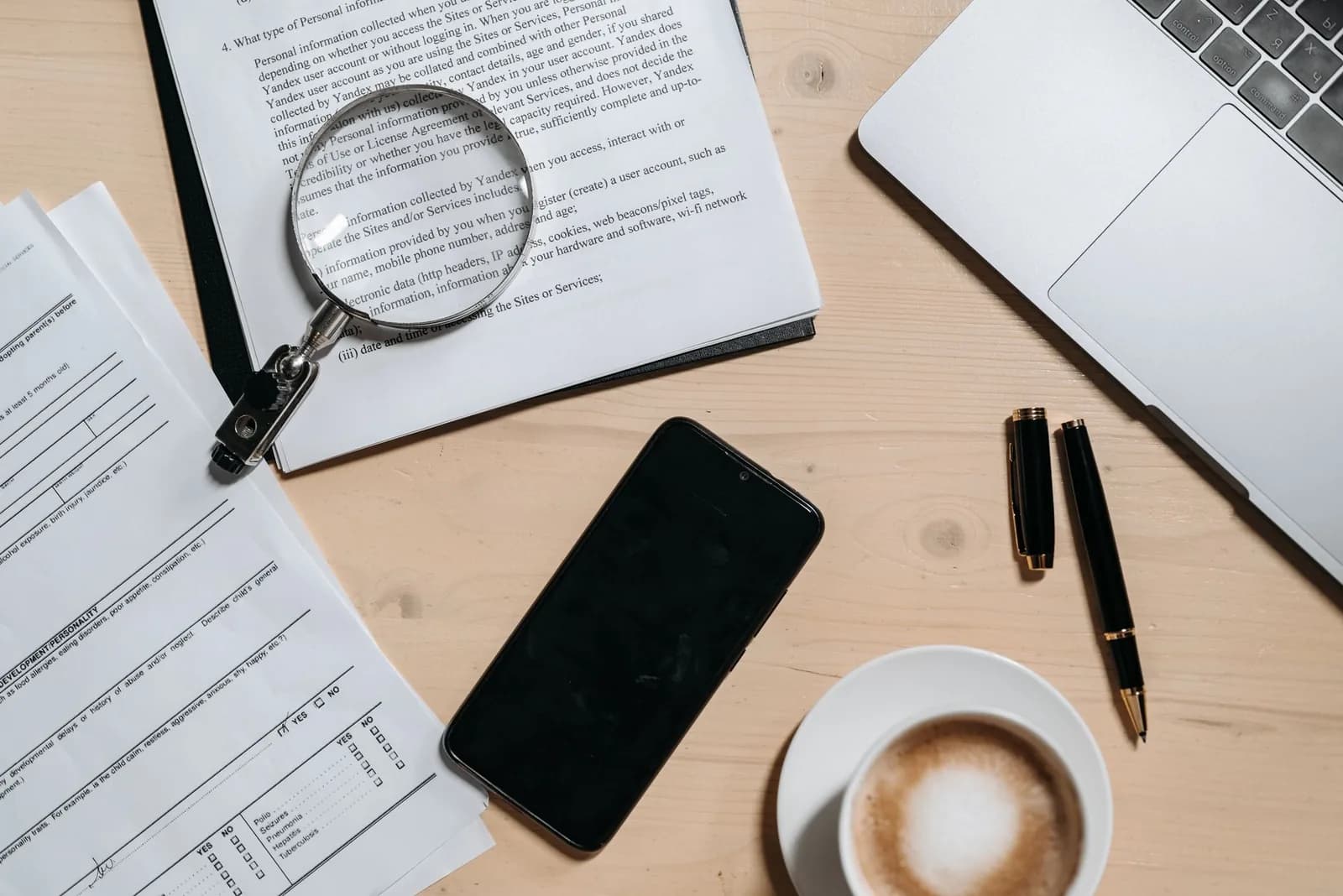 Overhead view of a magnifying glass resting on printed documents beside a smartphone, a fountain pen, a cup of coffee and a laptop on a pale wooden desk