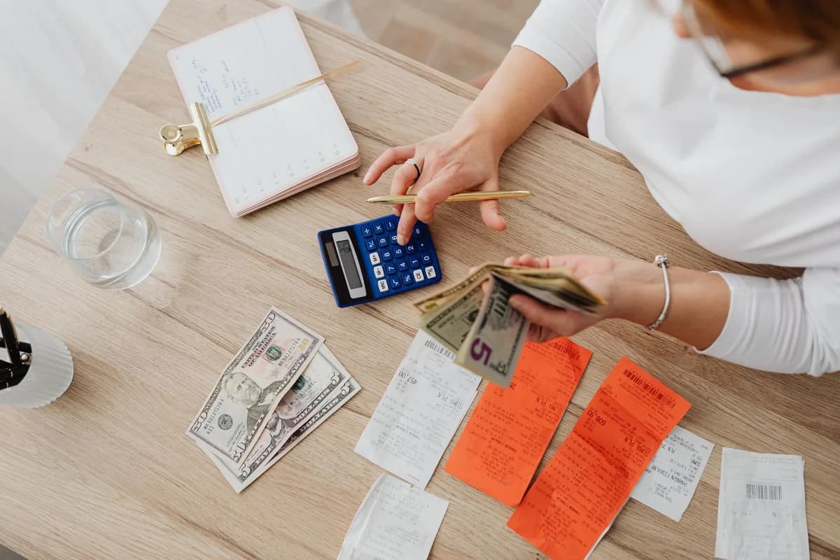 Overhead view of a light wood desk with a blue calculator, a few dollar bills, orange and white paper receipts and a notebook