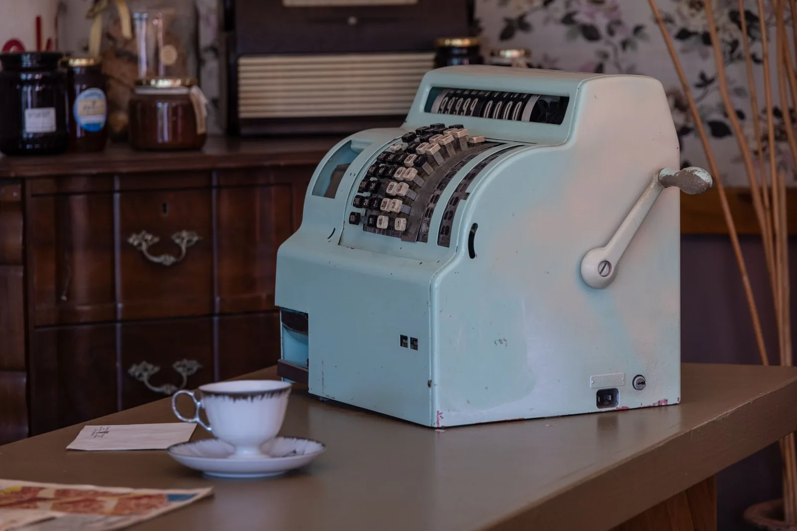 A pale green vintage cash register with a hand crank on a shop counter beside a white teacup and saucer
