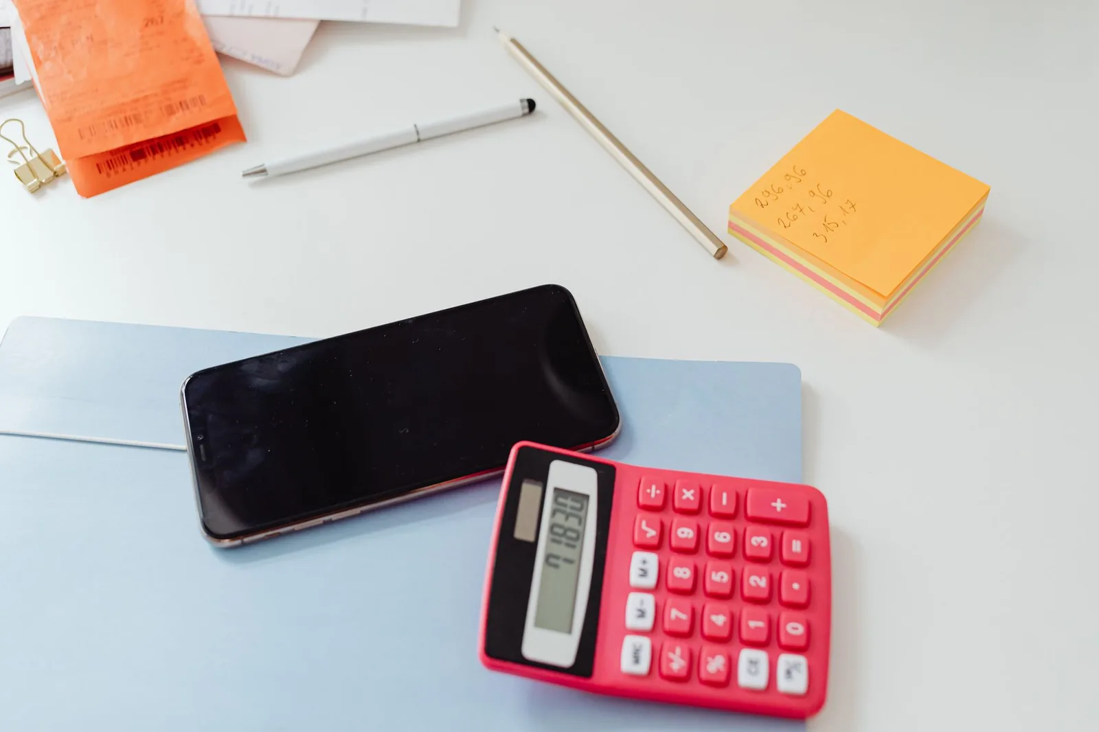 A calculator, phone, pay stub and a notepad with handwritten totals spread across a desk