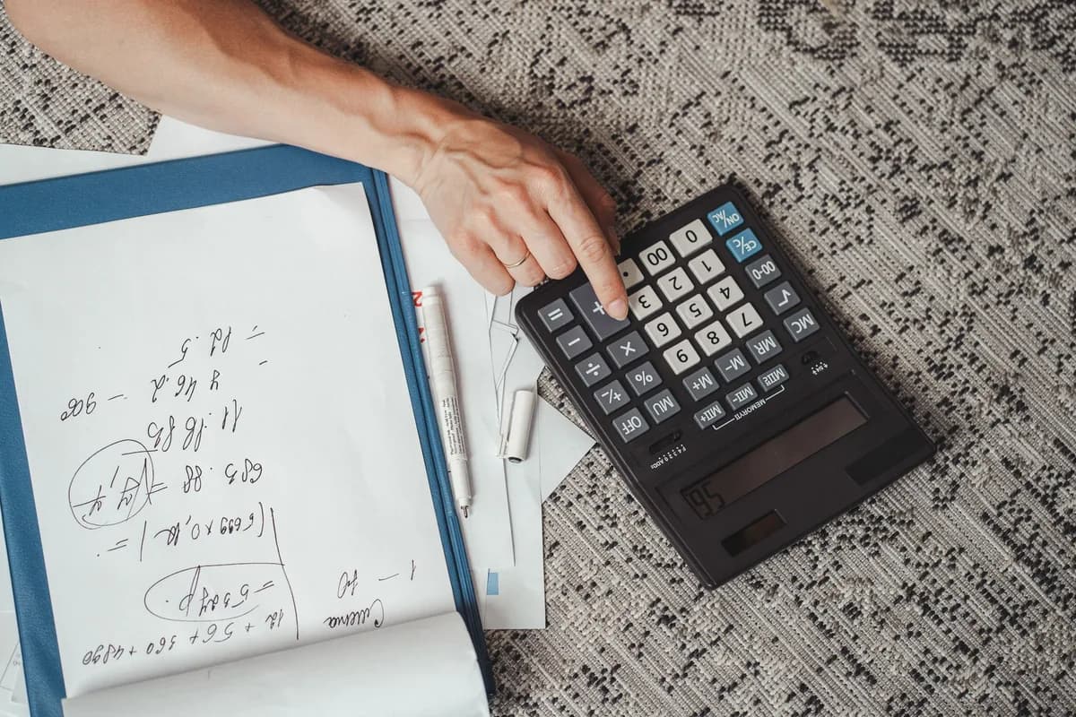 A hand pressing keys on a large black calculator beside a folder of handwritten sums on a textured tablecloth