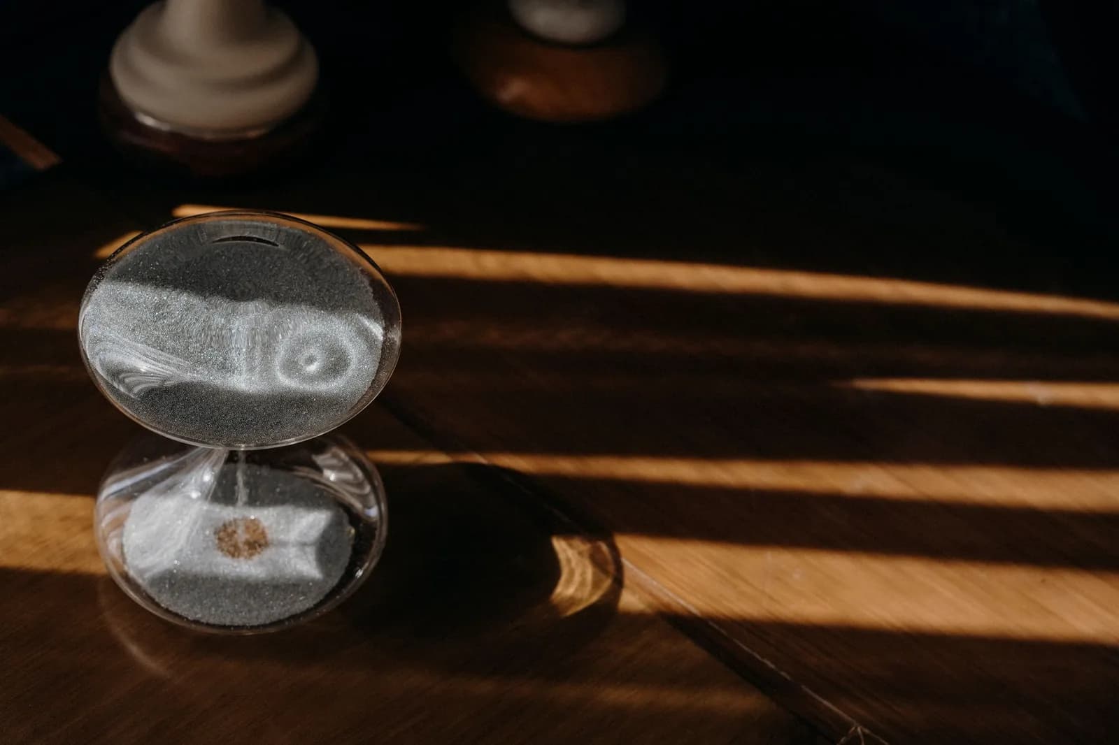 A glass hourglass of pale sand on a dark wooden table, crossed by bands of low afternoon sunlight
