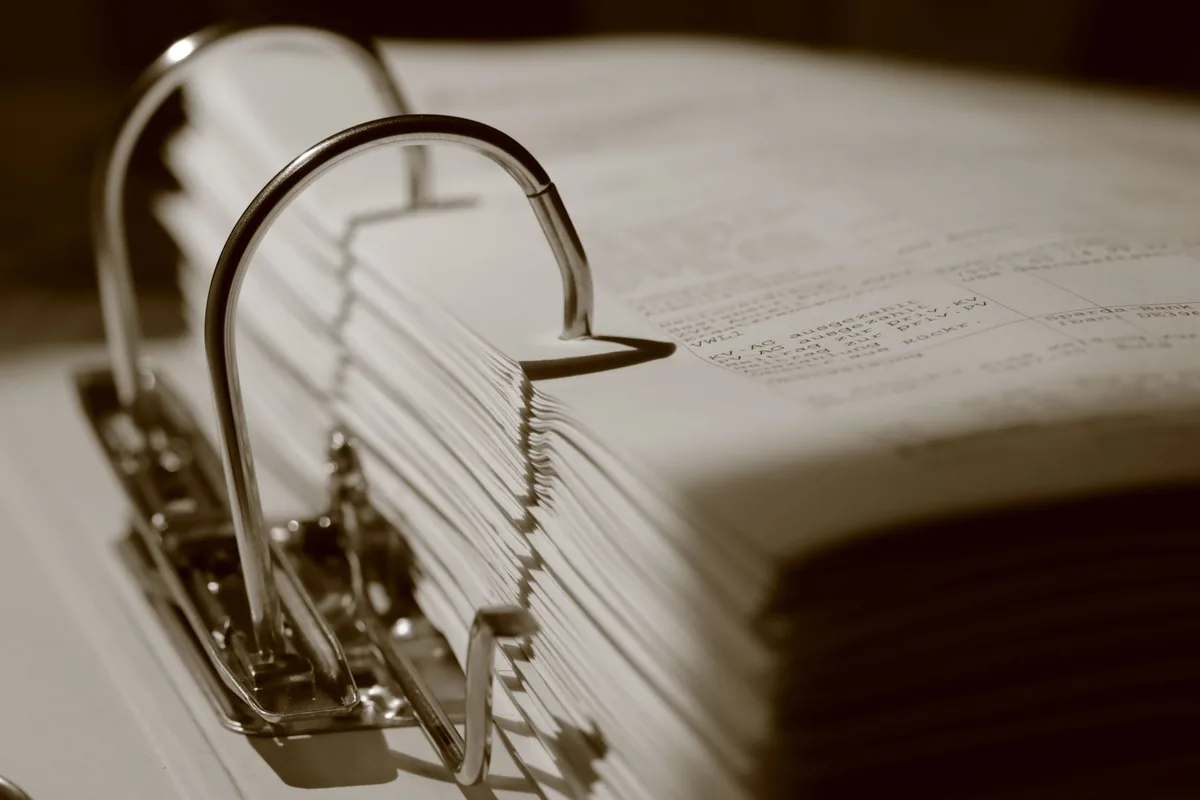 A sepia close-up of an open ring binder packed with typed pages, its metal rings curving over the edge of the stack