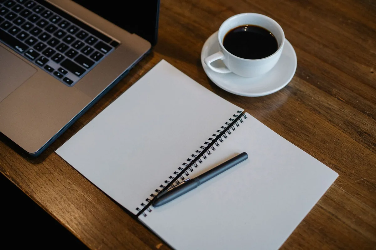 A blank spiral notepad with a black pen on a wooden table beside a cup of black coffee and an open laptop