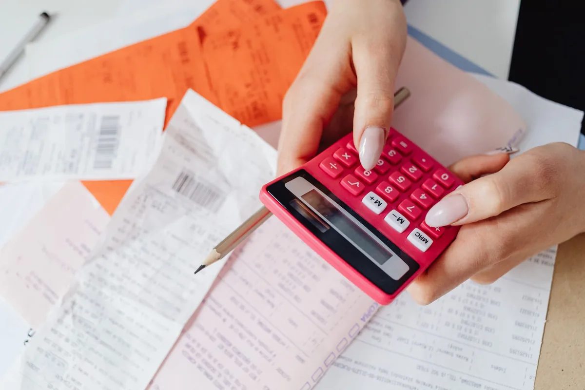 Two hands working a pink calculator over a spread of paper receipts, orange forms and a pencil on a desk