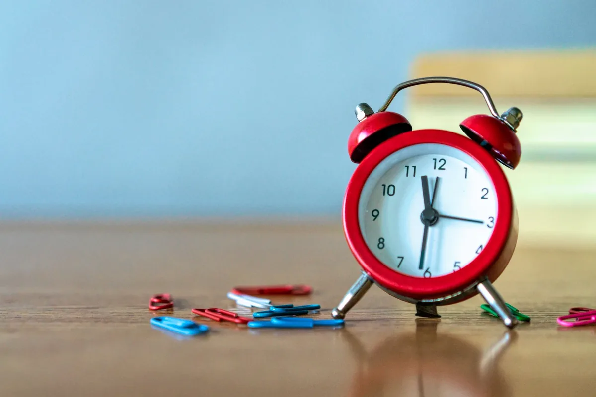 A small red twin-bell alarm clock standing on a wooden desk beside a scatter of coloured paper clips
