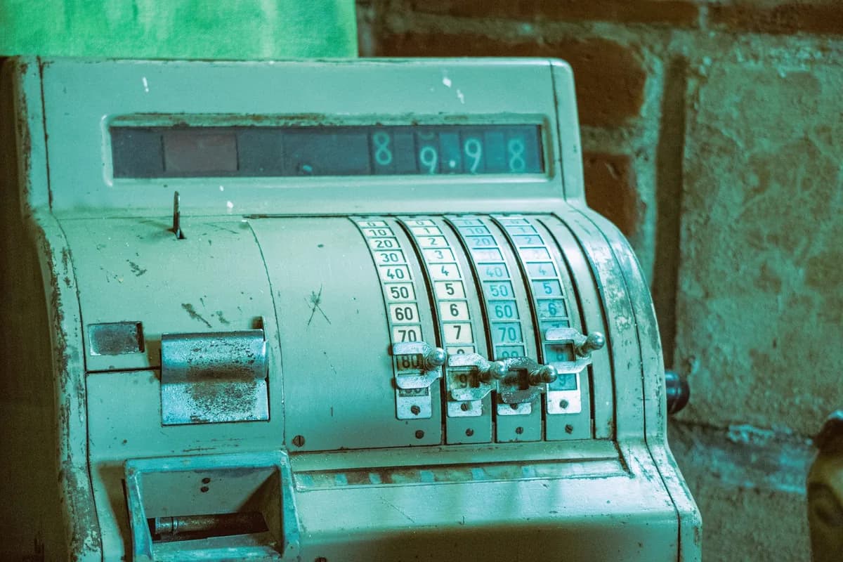 A worn pale-green vintage cash register with rows of numbered levers and a row of number wheels in its display, standing in front of a rough brick wall
