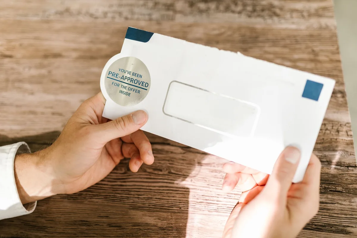 A person's hands holding a white window envelope stamped with a You have been pre-approved for the offer inside sticker over a wooden table