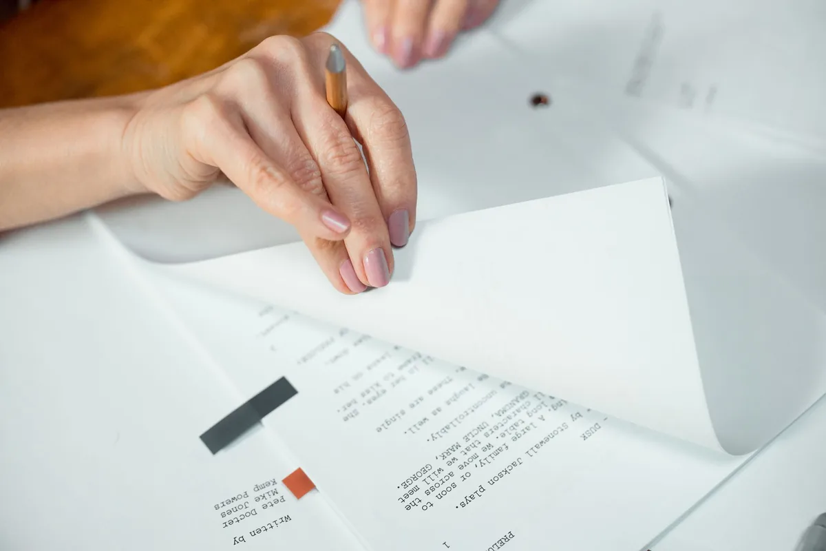 Close-up of a person's hands turning the pages of a printed multi-page document on a white desk, a pencil held between the fingers
