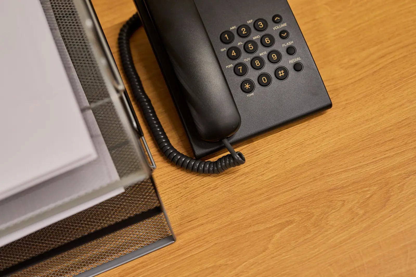 A classic black push-button desk telephone beside a wire document tray holding white papers on a wood-veneer office desk