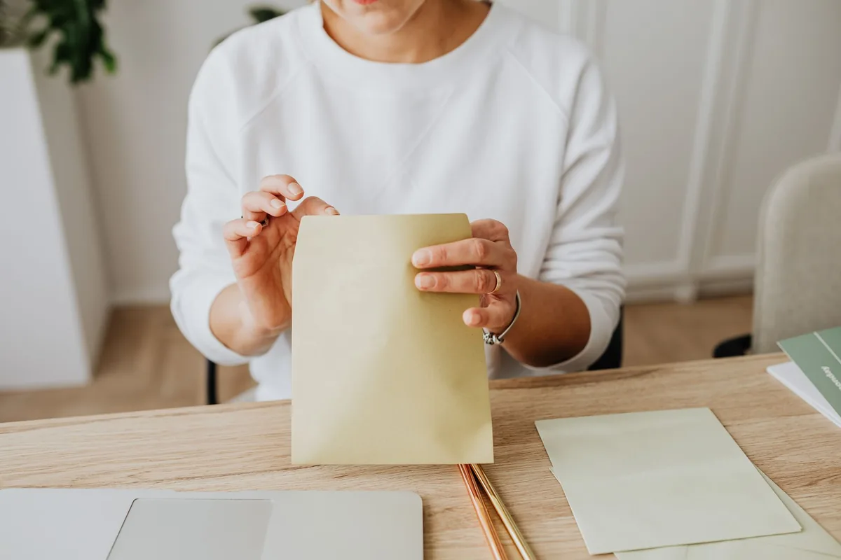 Hands holding a plain cream envelope over a light wooden desk with sheets of pale green paper and a pencil