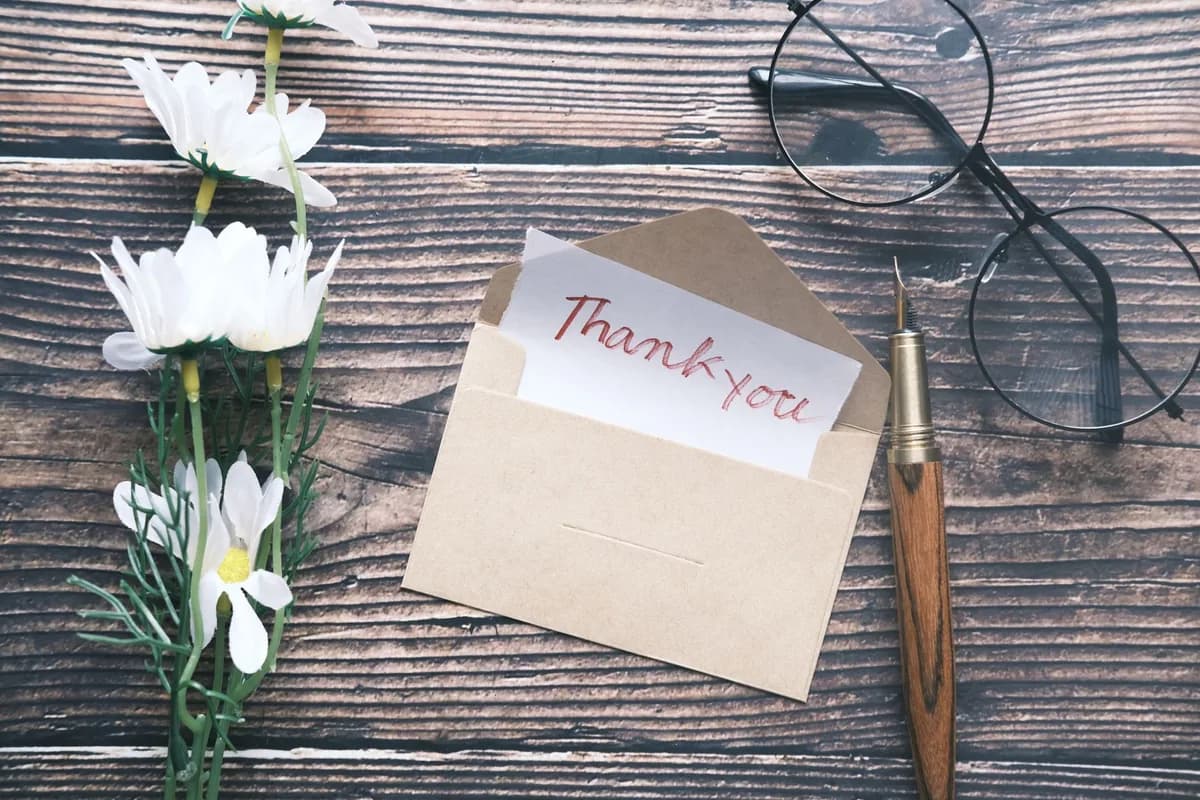 A handwritten thank-you note tucked into a kraft envelope on a dark wooden table, beside white daisies, reading glasses and a fountain pen