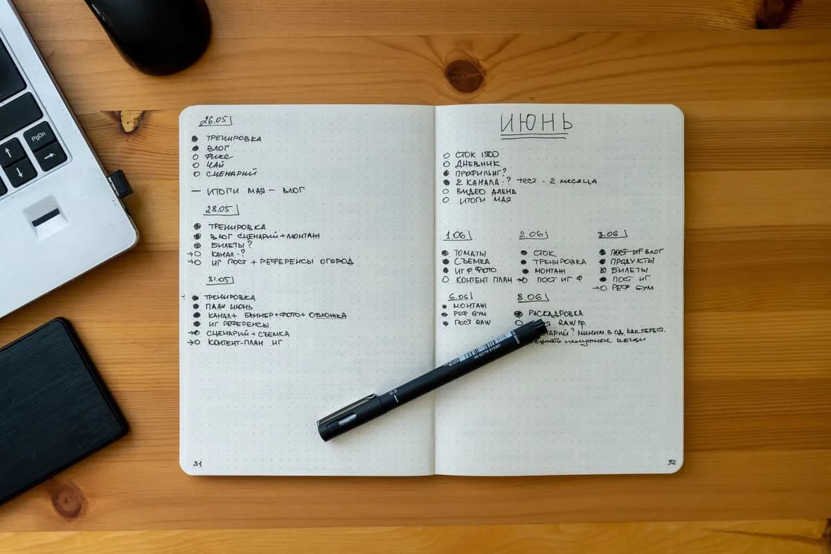 An open spiral notebook filled with handwritten daily entries and to-do lists, resting on a wooden desk beside a laptop, a computer mouse, and a pen