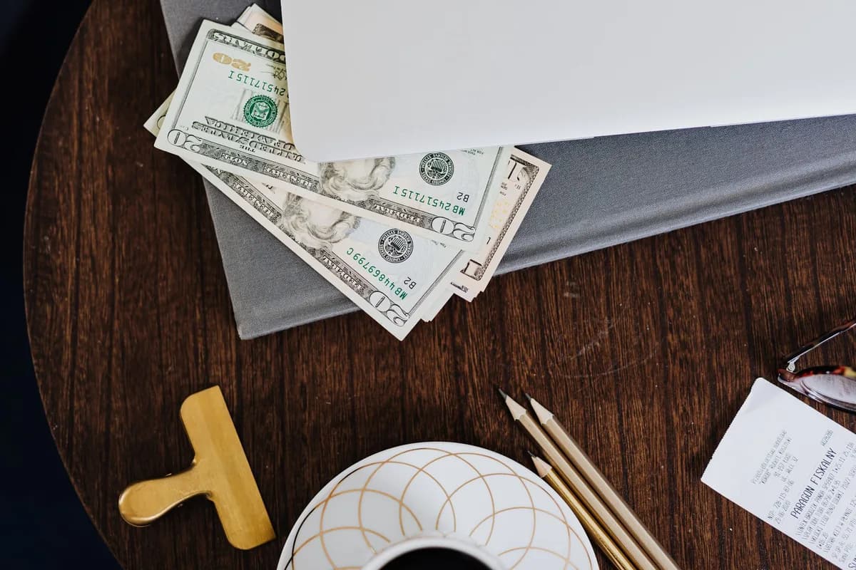 Several folded dollar bills tucked under a closed laptop on a round dark wooden table, with pencils, a brass clip and a coffee cup beside it