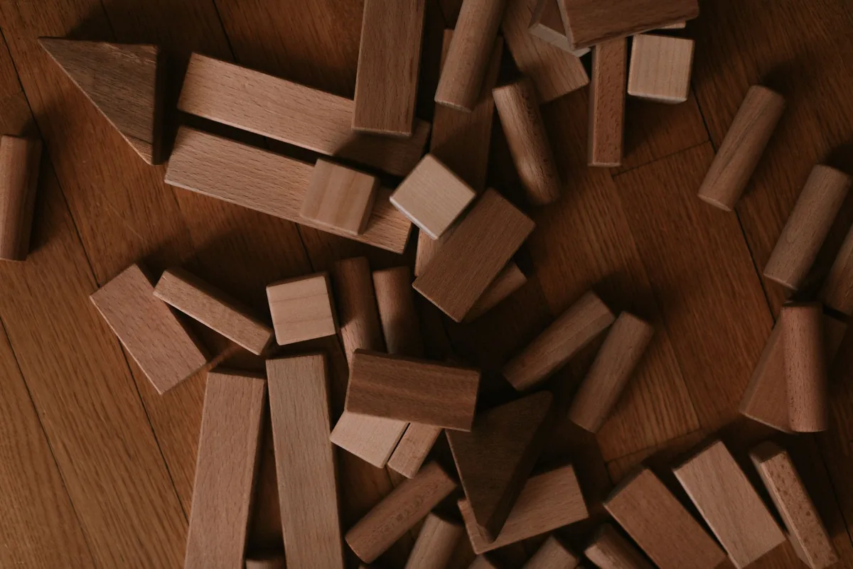 Plain wooden building blocks in assorted shapes scattered across a dark wooden floor, seen from above