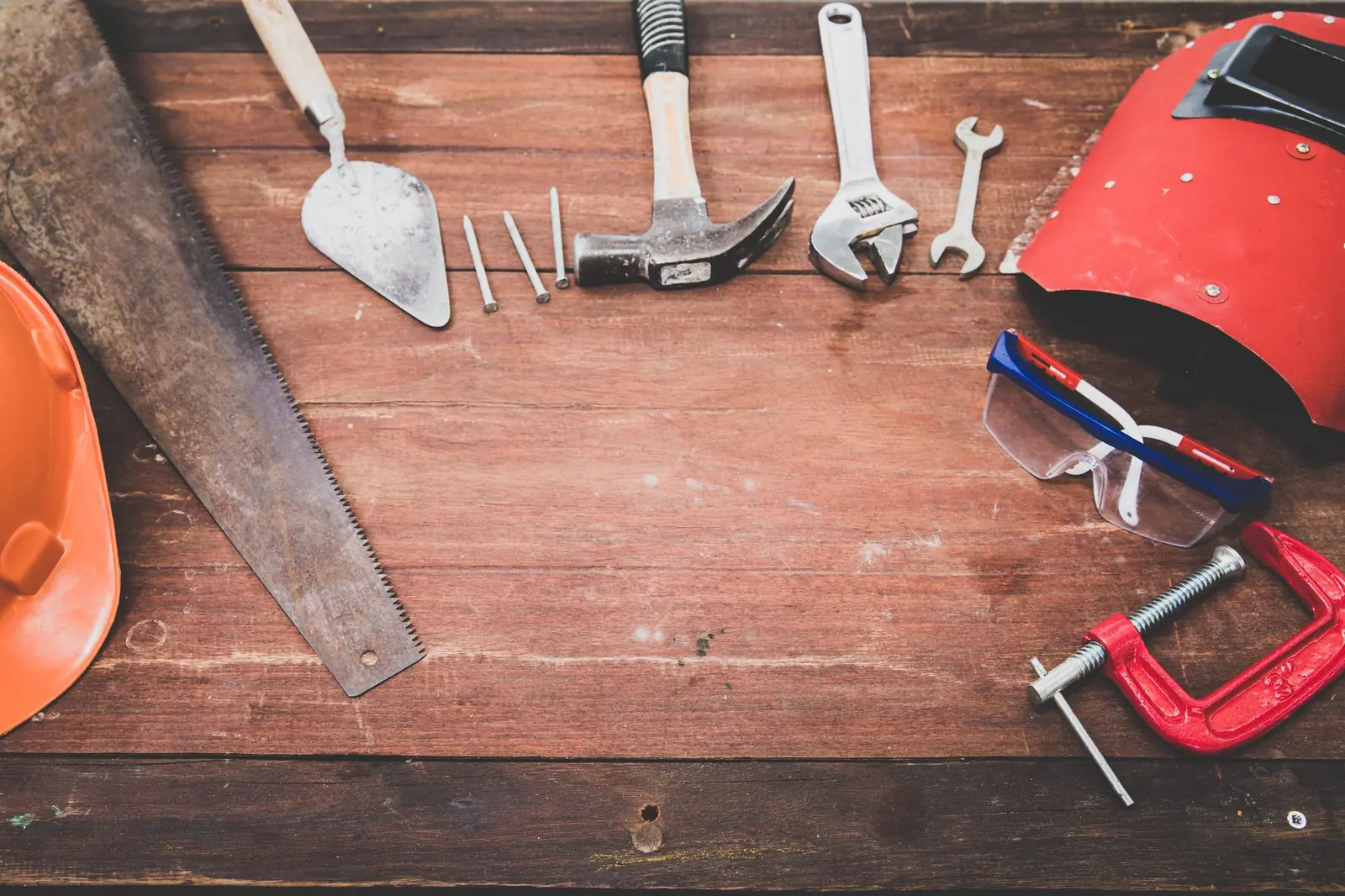 Hand tools laid out on a worn wooden table: a hand saw, trowel, hammer, wrenches, nails, safety glasses, a clamp and an orange hard hat