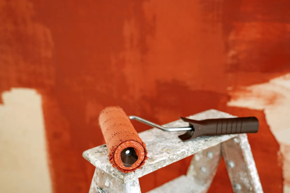 A paint roller with rust-red paint resting on a paint-spattered stepladder in front of a half-painted wall