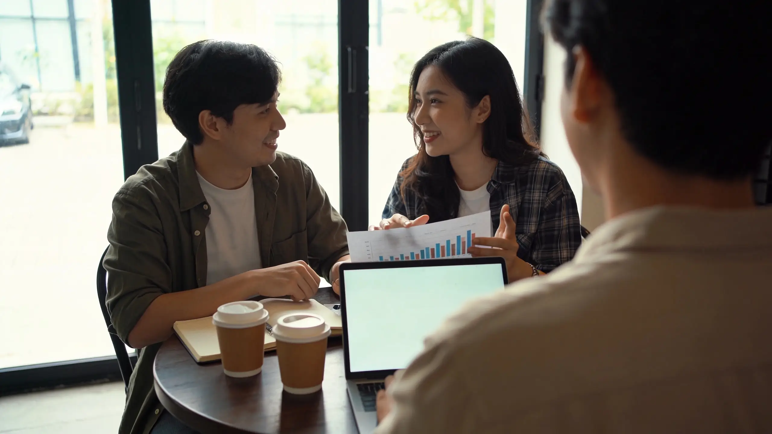 Two people at a small round cafe table with coffee, a laptop, and an open notebook, mid-conversation