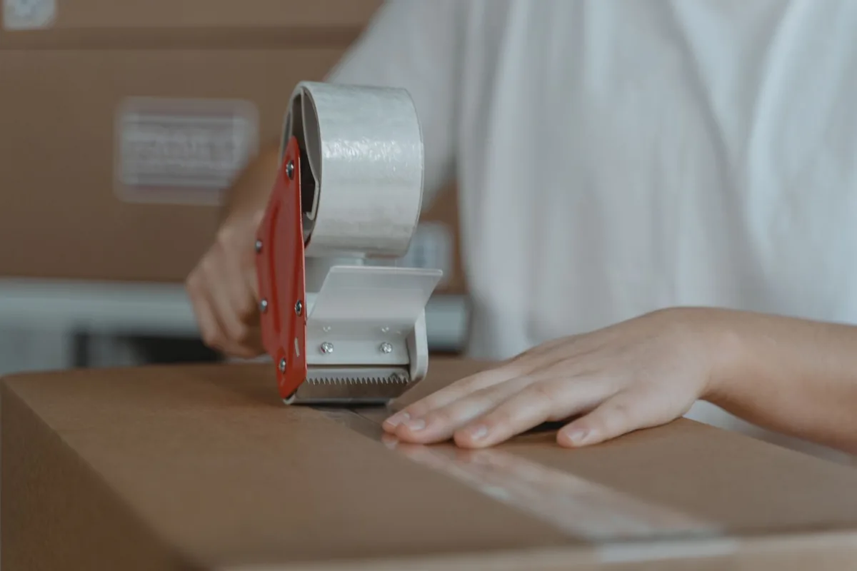 A pair of hands running a tape gun along the seam of a closed cardboard shipping box