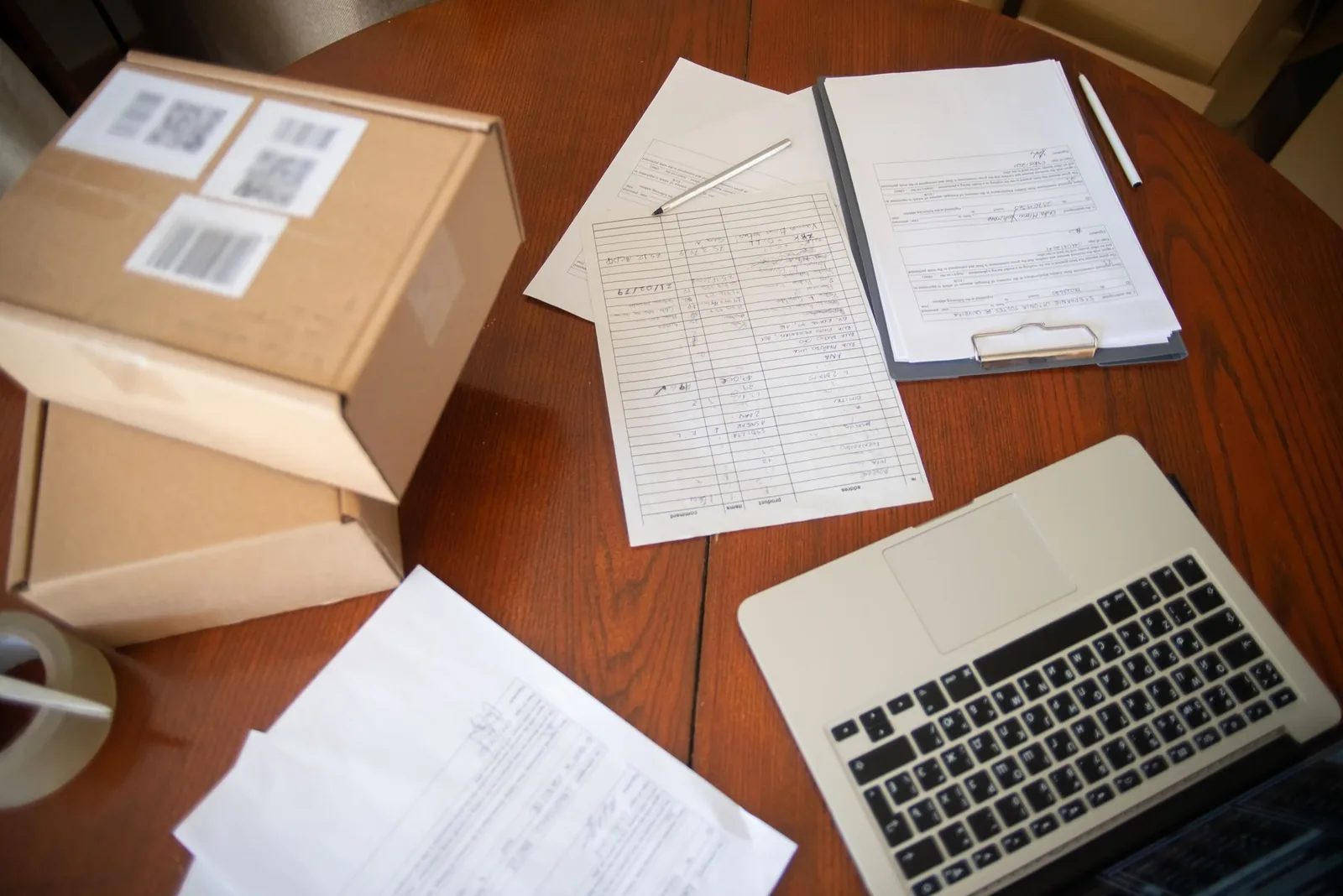 An overhead view of a wooden desk with labelled shipping boxes, a tape dispenser, printed order forms on a clipboard and an open laptop