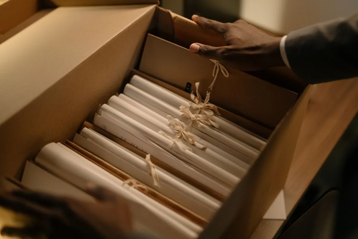 A person's hands sorting through tied bundles of documents in a cardboard file box under warm light