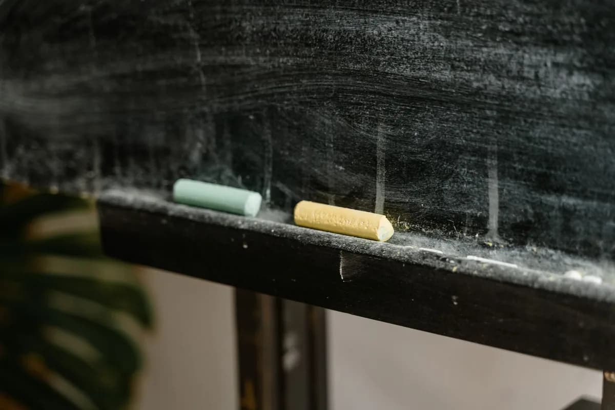 A green and a yellow stick of chalk resting on the dusty wooden ledge of a wiped blackboard