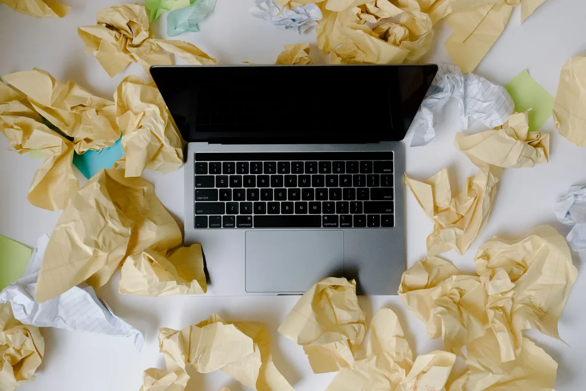 Overhead view of an open laptop on a white surface surrounded by crumpled sheets of yellow, white and green paper