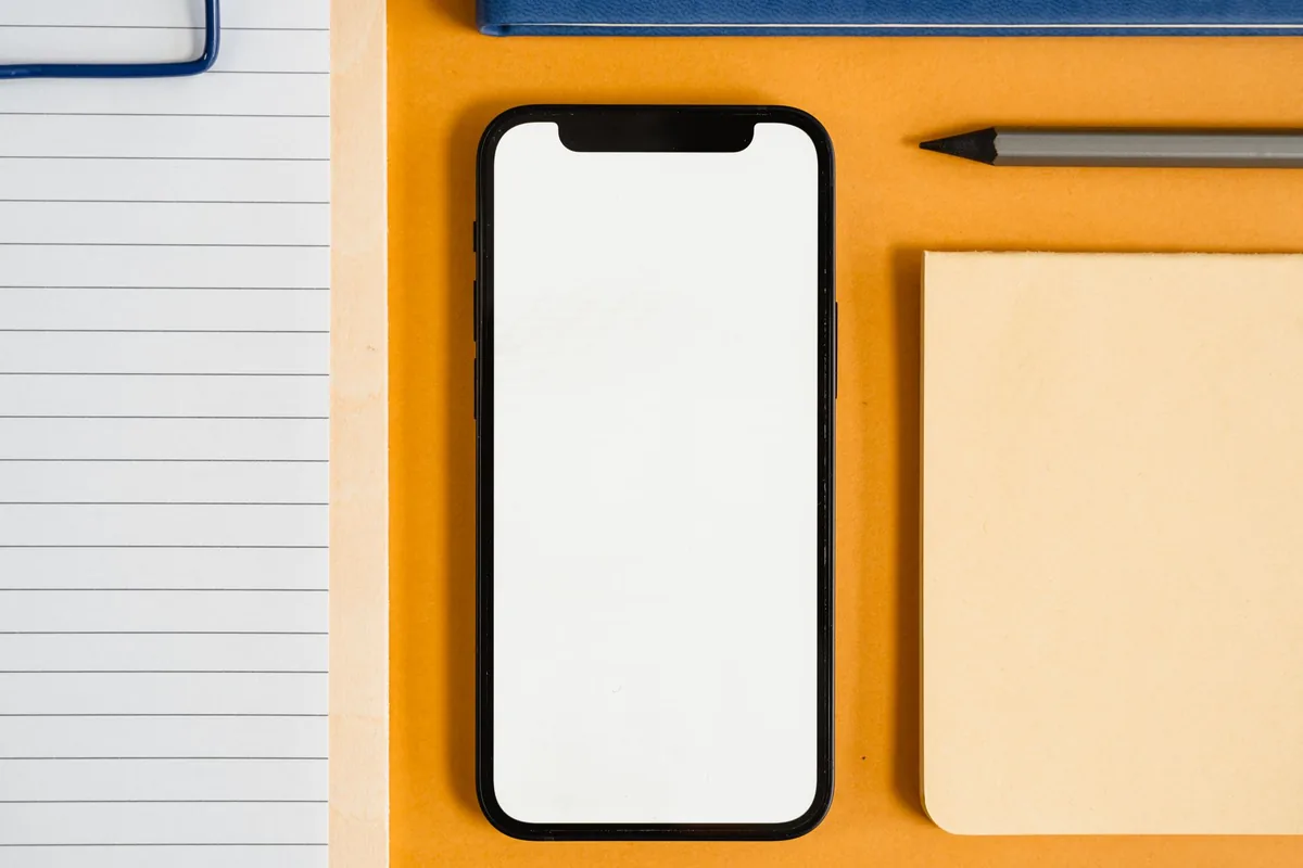 A phone with a blank screen resting on an orange desk beside a notepad and pencil